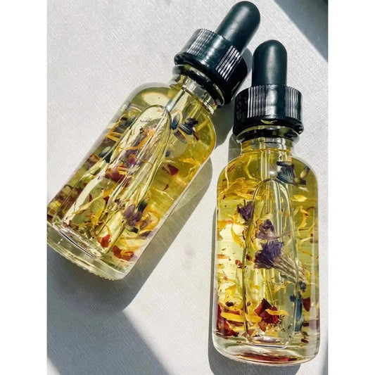 Two clear glass dropper bottles of golden ritual oil infused with dried flower petals on a white surface.
