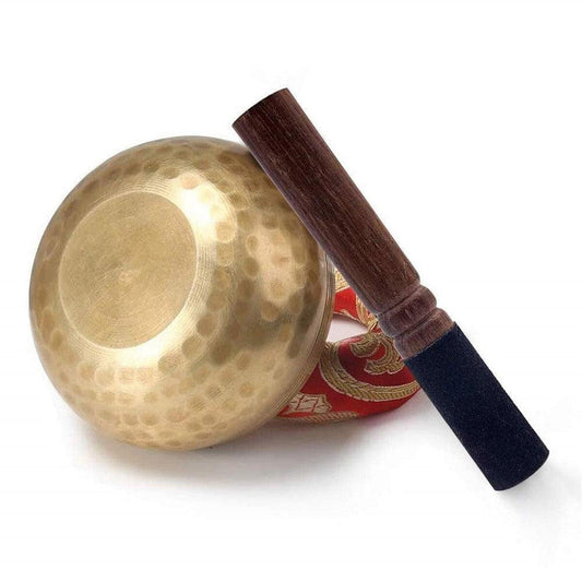 Handcrafted hammered copper Tibetan singing bowl with a wooden striker and decorative red cushion on white background.