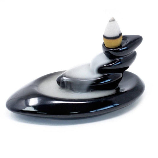 Black ceramic backflow incense burner with a stacked stone design and cascading white smoke on a white background.