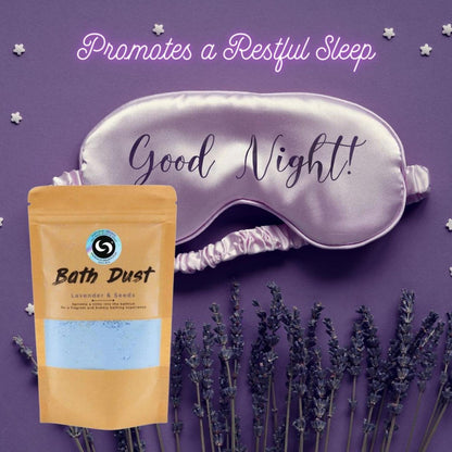 Lavender and Seeds bath dust pouch next to a satin sleep mask on a purple background with dried lavender sprigs.