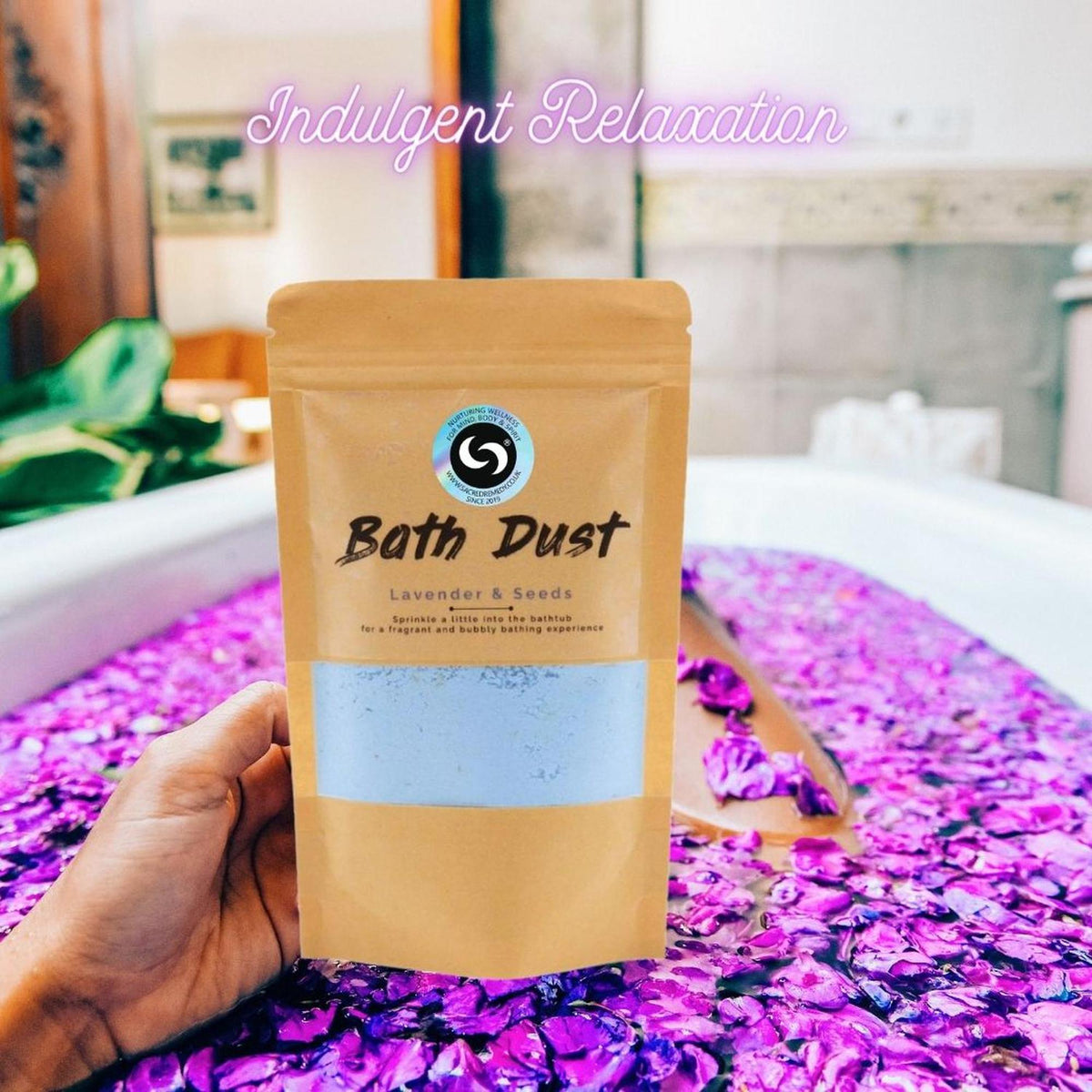 Hand holding a pouch of Lavender & Seeds bath dust in front of a bathtub filled with purple flower petals and water.