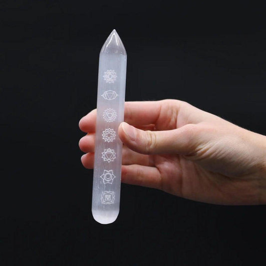 Hand holding a translucent, cylindrical selenite crystal wand engraved with seven white chakra symbols.