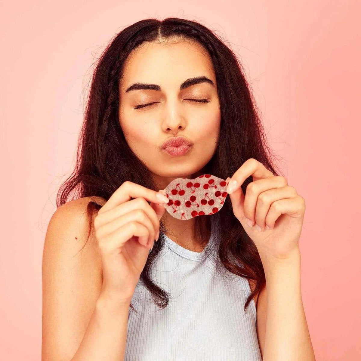 Woman holding a translucent hydrogel lip mask with a red cherry pattern against a soft pink background.