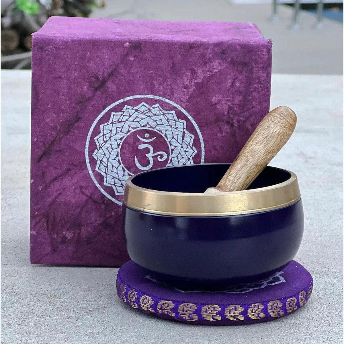 Deep blue singing bowl with brass rim, wooden striker, and purple Om-symbol gift box on a matching embroidered cushion.