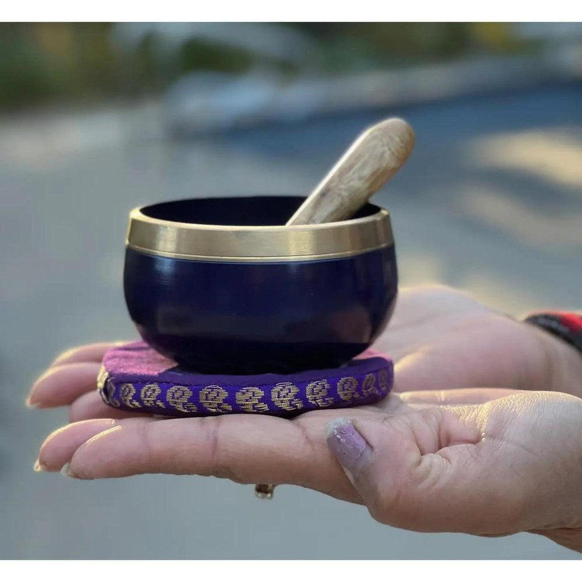 Deep purple Crown Chakra singing bowl with brass rim, wooden striker, and patterned cushion held in hands.