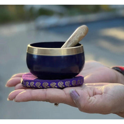 Deep purple Crown Chakra singing bowl with brass rim, wooden striker, and patterned cushion held in hands.