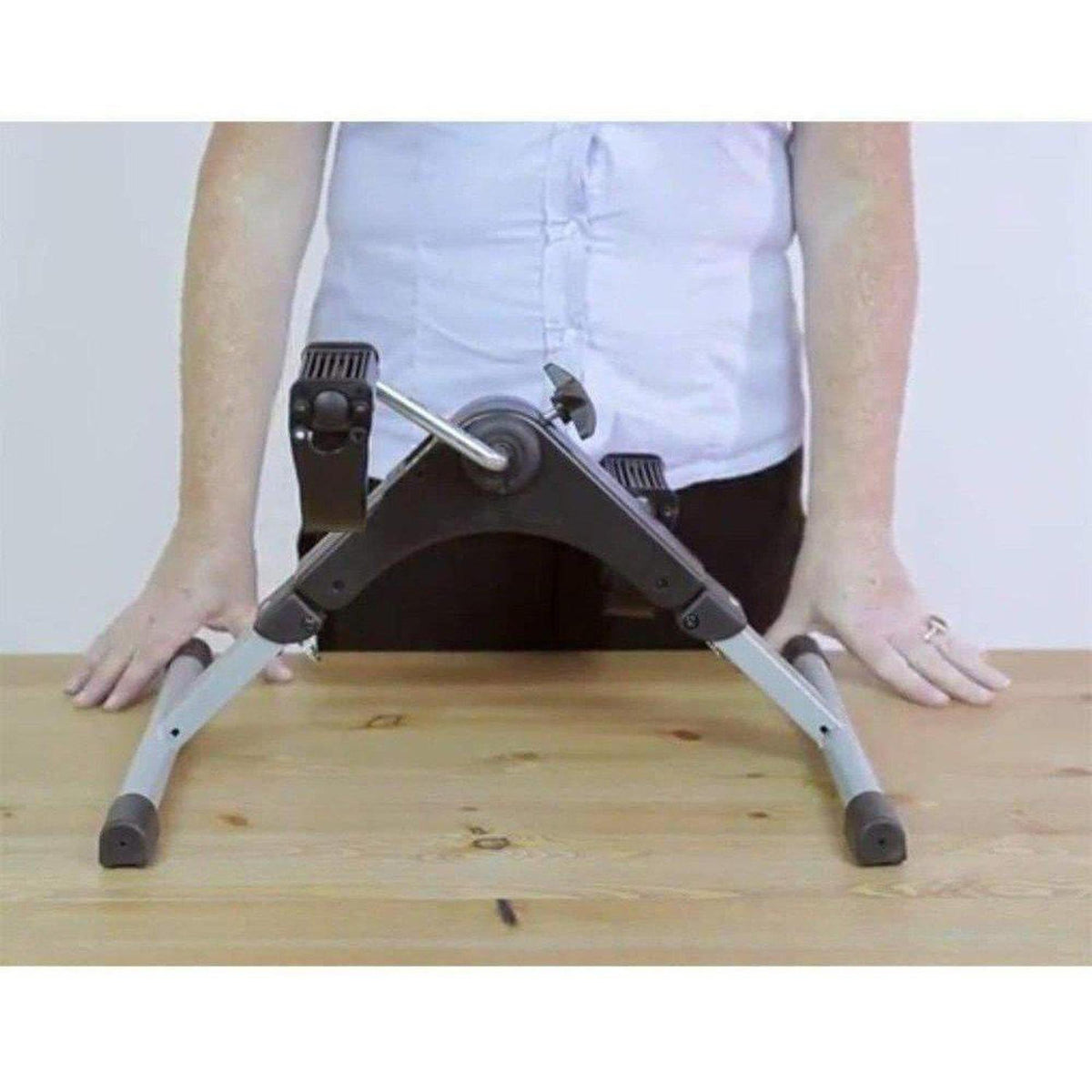 Black and silver portable pedal exerciser with adjustable resistance knob placed on a wooden table for arm exercise.