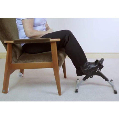 Person using a black digital pedal exerciser with adjustable resistance while seated in a chair for low-impact leg exercise.