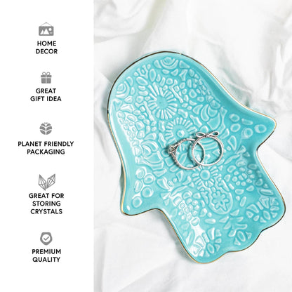 Turquoise Hamsa hand jewellery dish with intricate embossed patterns and gold-trimmed edges holding two silver rings.