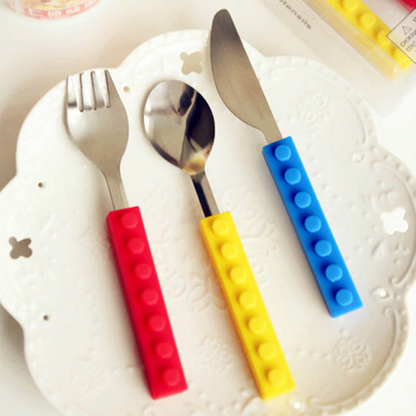Three-piece stainless steel kids' cutlery set with red, yellow, and blue silicone building block handles on a white plate.