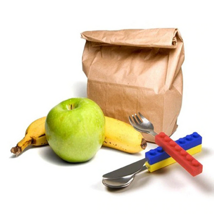 Stacking kids' cutlery set with colorful silicone building block handles, stainless steel utensils, fruit, and a lunch bag.