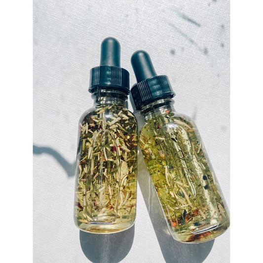 Two clear glass dropper bottles filled with ritual oil, dried botanicals, and herbs on a white textured background.