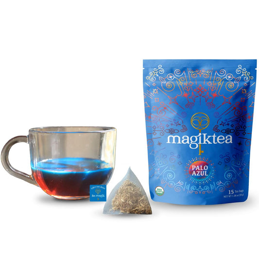 Glass cup of glowing blue Palo Azul tea next to a Magiktea pouch and a pyramid tea bag on a white background.