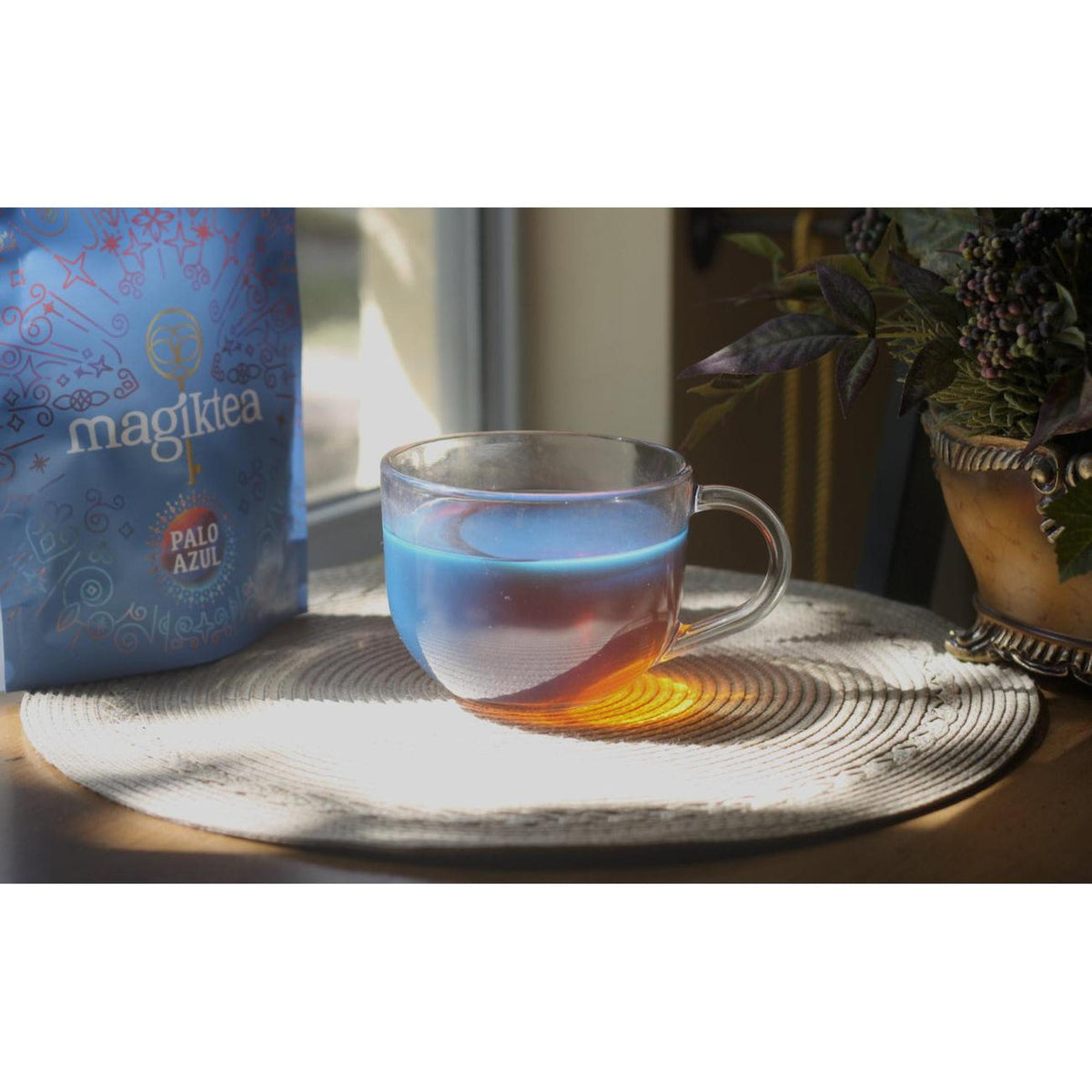 Glass mug filled with fluorescent blue Palo Azul tea next to a Magiktea branded pouch on a woven placemat.