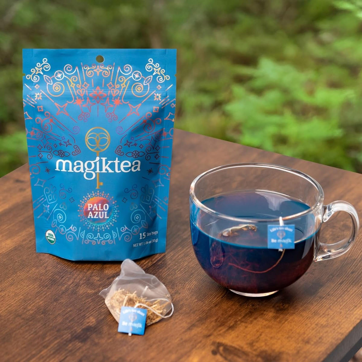 Magiktea Palo Azul pouch next to a clear glass mug filled with glowing blue herbal tea on a wooden table.