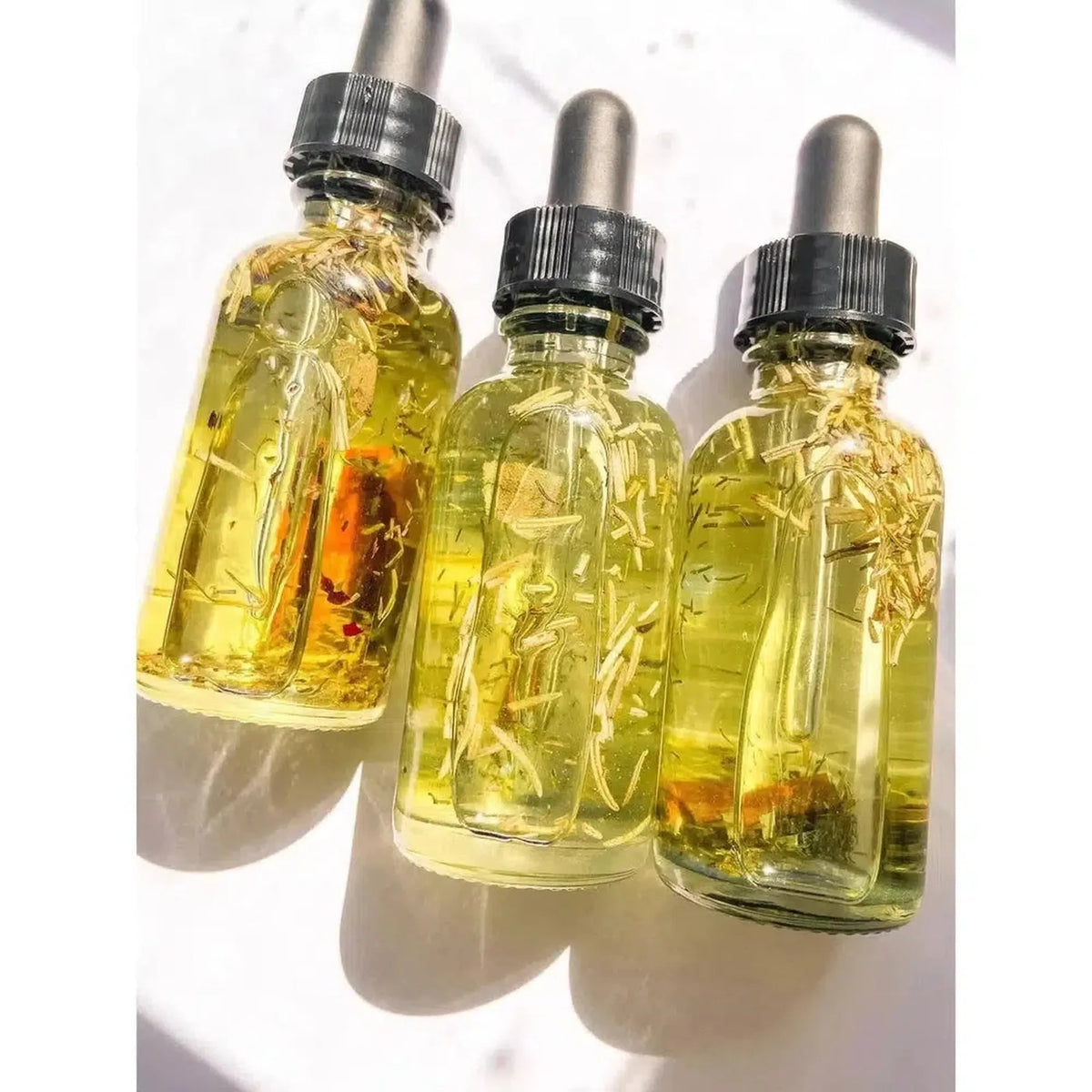 Three glass dropper bottles filled with golden ritual oil, botanical herbs, and shredded currency on a white surface.