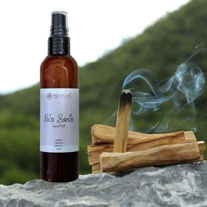 Amber spray bottle of Palo Santo smudge mist next to a burning wooden stick on a stone surface outdoors.