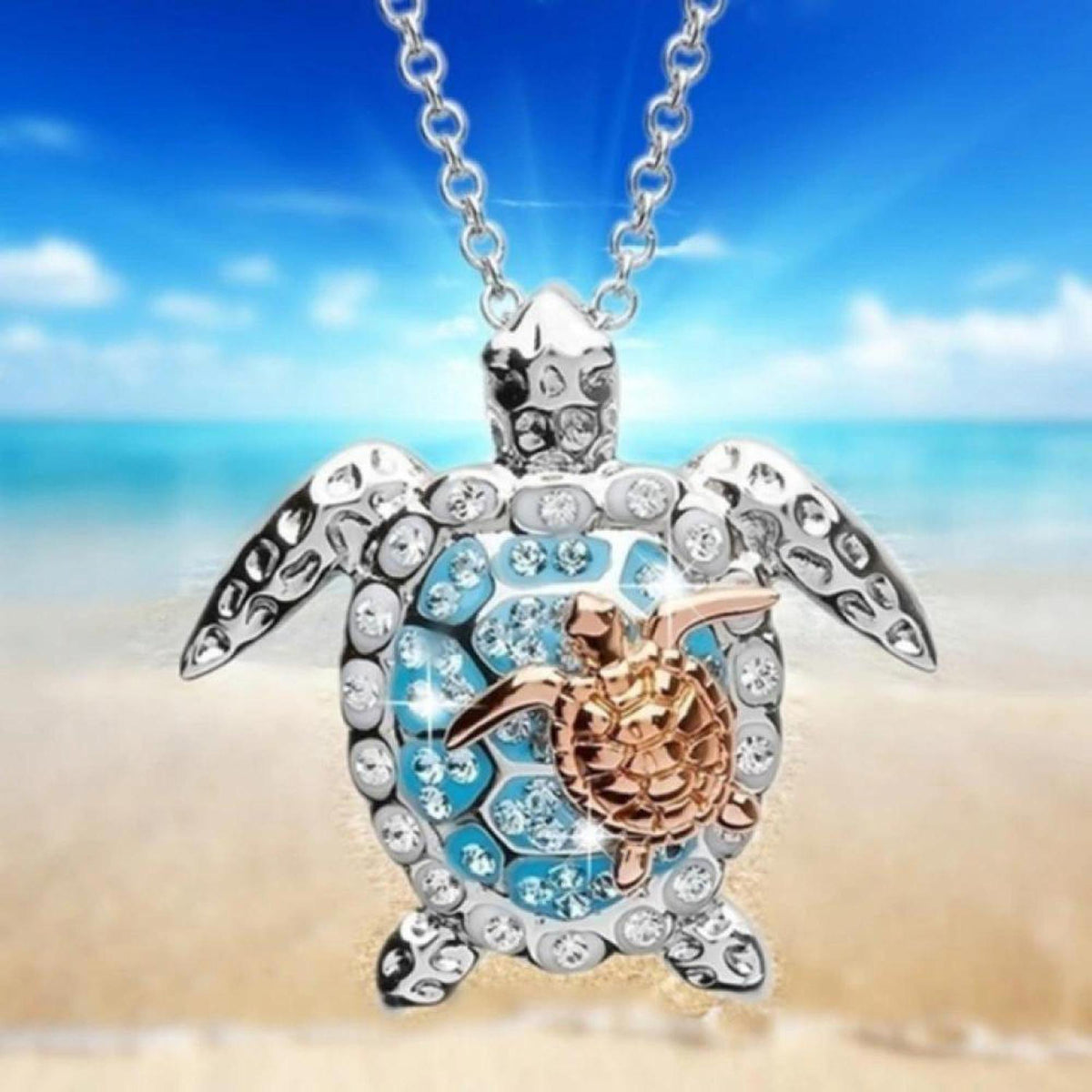 Sterling silver sea turtle necklace with a rose gold baby turtle, blue enamel, and sparkling zirconia on a beach background.