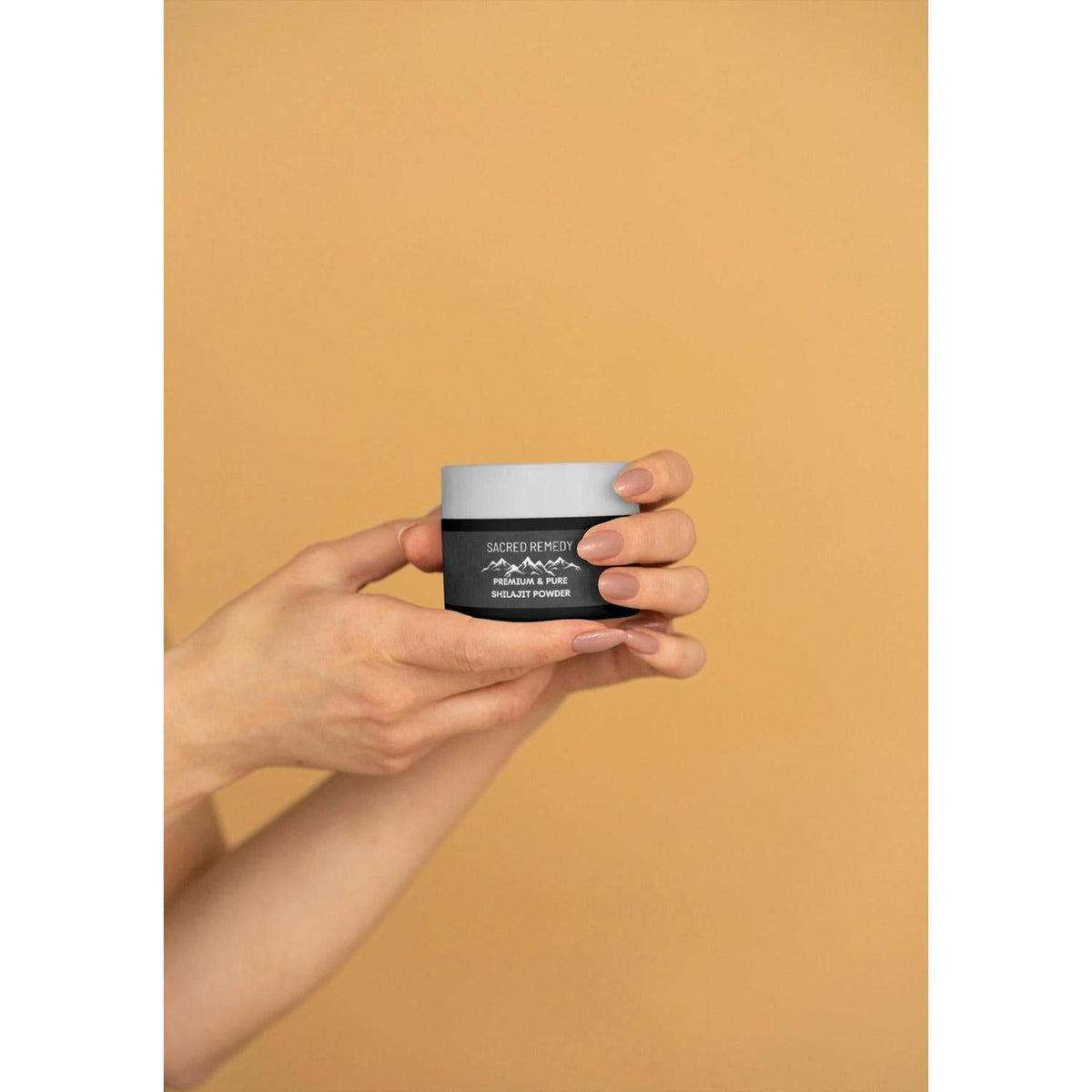 Hands holding a small black and white jar of Sacred Remedy premium Himalayan Shilajit powder on a tan background.
