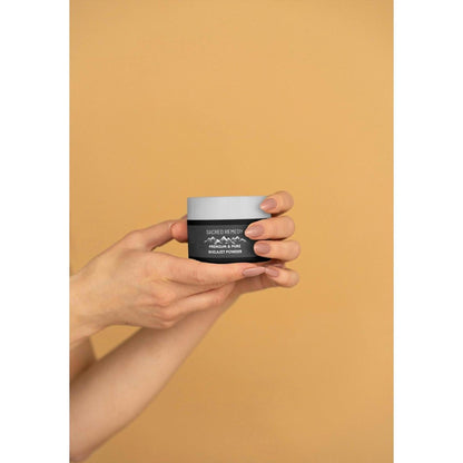 Hands holding a small black and white jar of Sacred Remedy premium Himalayan Shilajit powder on a tan background.