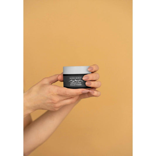 Hands holding a small black and white jar of Sacred Remedy premium Himalayan Shilajit powder on a tan background.