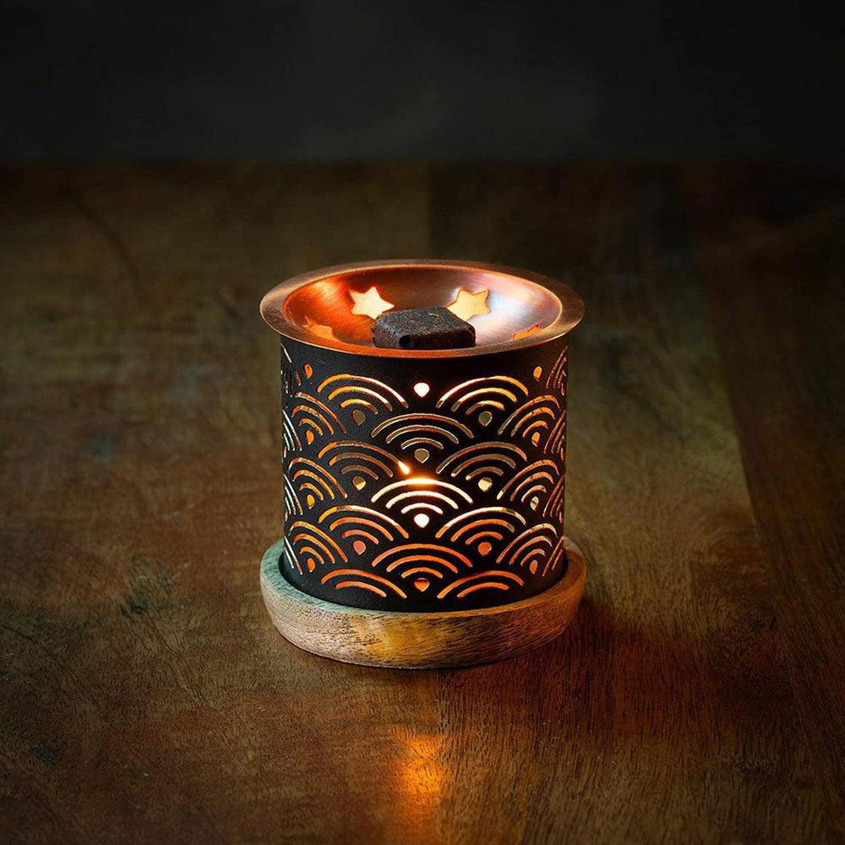 Black metal incense burner with wave cutouts, copper top, and wooden base, lit by a tea light candle on a wooden table.