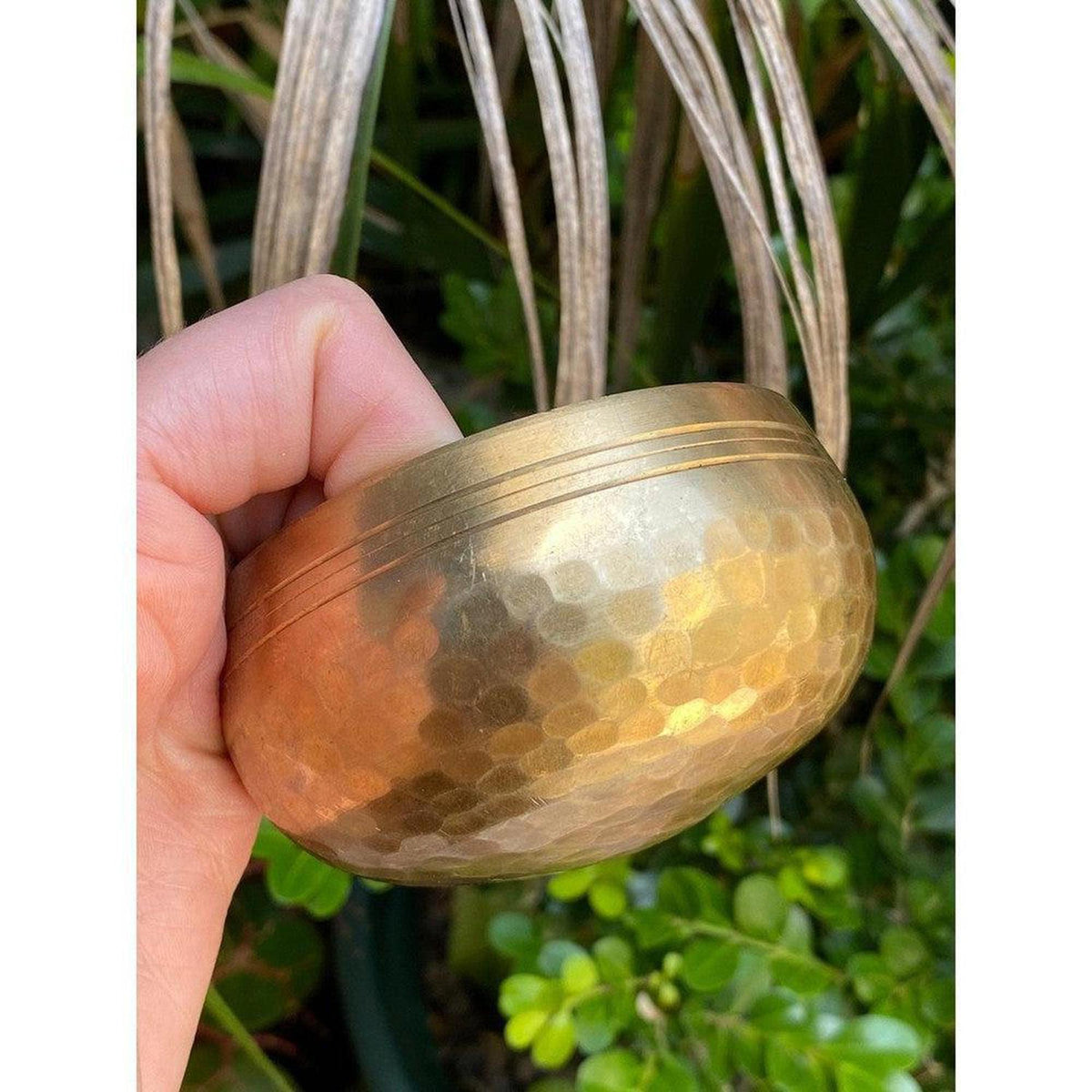 Handcrafted copper Tibetan singing bowl with a hammered texture and etched rim held against a lush green garden background.