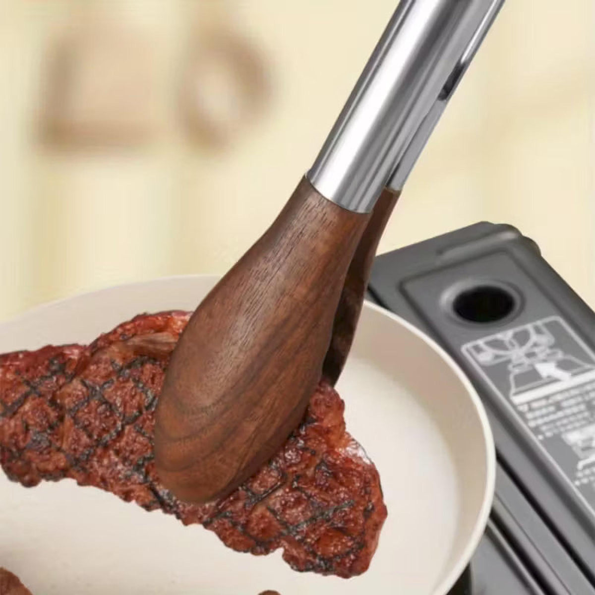 Stainless steel kitchen tongs with natural walnut wood heads lifting a seared steak from a non-stick pan.